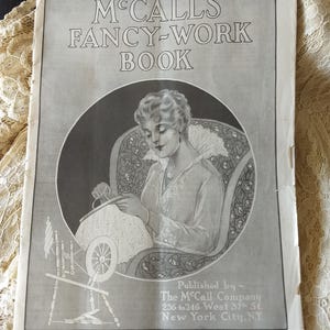 May include: A vintage "McCall's Fancy-Work Book" cover. The monochrome illustration shows a woman seated, doing needlework. The book's title is displayed, along with the publisher's details: The McCall Company, New York City.