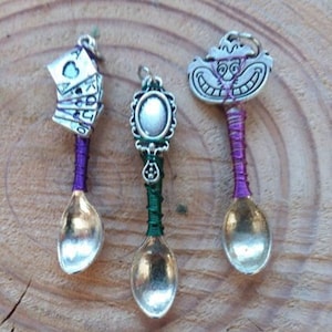 Puede incluir: Tres cucharas decorativas con cuencos plateados y mangos envueltos. Una cuchara presenta una pila de naipes, otra tiene un marco ovalado y la tercera tiene una cara de Gato de Cheshire. Los mangos están envueltos en alambre morado y verde.