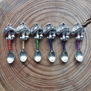 Puede incluir: Seis cucharas de plata con tapas en forma de hongo y mangos de cuerda de colores. Las cucharas están dispuestas en fila sobre una superficie de madera.