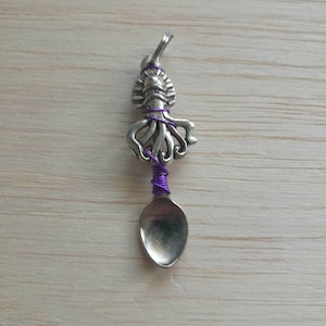 Puede incluir: Colgante de cuchara plateada con un diseño detallado de pulpo. El pulpo está envuelto con alambre morado, agregando un toque de color. Una pieza pequeña y única, perfecta para joyería o proyectos de manualidades.