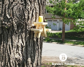 May include: A miniature wooden picnic table with a yellow corn cob on top, attached to a tree trunk.