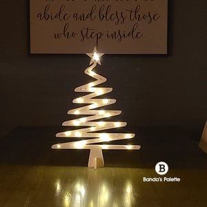 May include: A white wooden Christmas tree with a star on top and lights strung around it. The tree is sitting on a table in front of a sign that reads "In our home let love abide and bless those who step inside".
