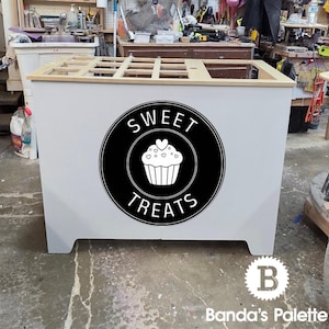 May include: A white, rectangular sweet treats display stand with a black circular logo featuring a cupcake and the words "SWEET TREATS". The stand is in a workshop setting, with tools and shelves visible in the background.