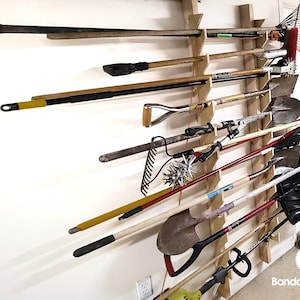 May include: A wooden wall-mounted storage rack with multiple slots for holding gardening tools. The rack is filled with various tools, including rakes, shovels, and a weed whacker.