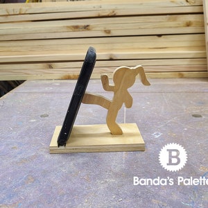 May include: A wooden phone stand featuring a girl kicking a phone. The stand is made of light brown wood and the girl is painted in a natural wood color. The phone is black and is being held up by the girl's leg. The stand is on a gray surface with the text "Banda's Palette" and a letter "B" in a circle.