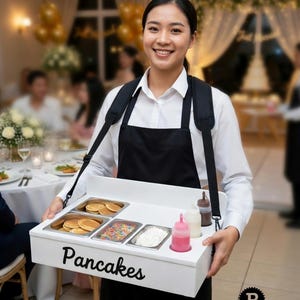May include: A white portable serving tray with black straps containing metal inserts filled with small pancakes, colorful sprinkles, and whipped cream, plus three squeeze bottles. The front of the tray features the word Pancakes in black text.