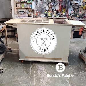 May include: A wooden charcuterie cart with a white worktop and a black and white logo that says "Charcuterie Cart". The cart has a built-in chopping board and a sink. The cart is on wheels and is in a workshop setting.