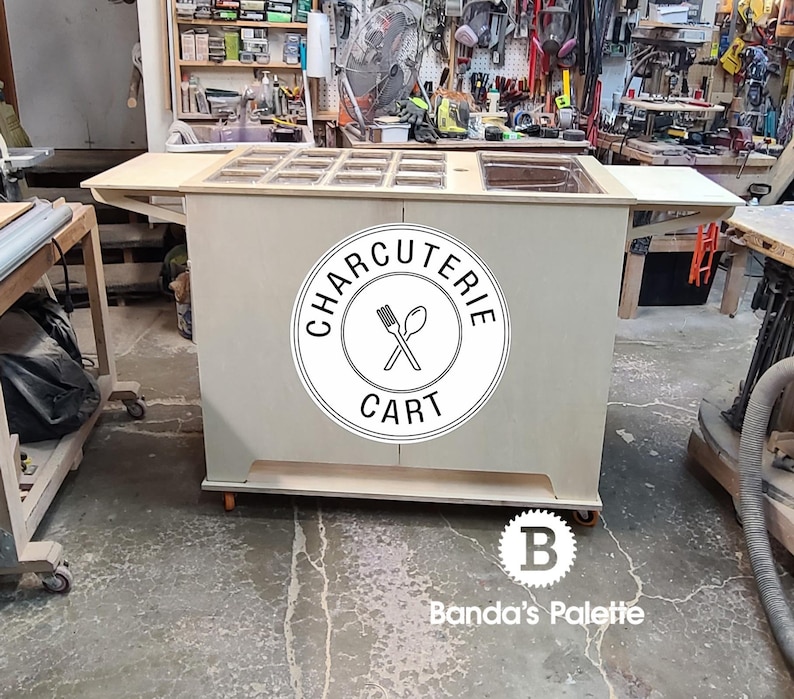 May include: A wooden charcuterie cart with a white worktop and a black and white logo that says "Charcuterie Cart". The cart has a built-in chopping board and a sink. The cart is on wheels and is in a workshop setting.