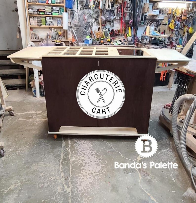 May include: A wooden charcuterie cart with a brown stained front panel and a white label that says "CHARCUTERIE CART" with a crossed fork and spoon icon. The cart has a fold-down serving shelf and a bottom shelf. The cart is on wheels and is in a workshop setting.