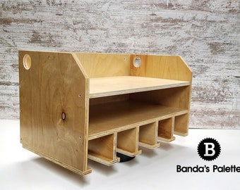 May include: A wooden wall-mounted organizer with a shelf and six compartments. The organizer is made of light-colored wood and has a natural finish. The compartments are designed to hold various items, such as tools or paintbrushes. The organizer is labeled "Banda's Palette".