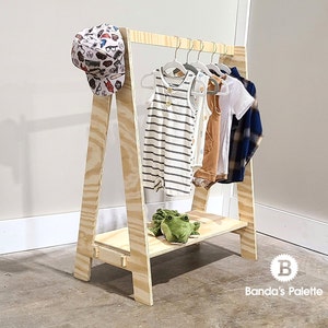 May include: A-frame wooden clothing rack with a shelf, holding several baby outfits on hangers. A patterned baseball cap hangs on the side. A green stuffed frog sits on the shelf. The rack is made of light-colored wood.