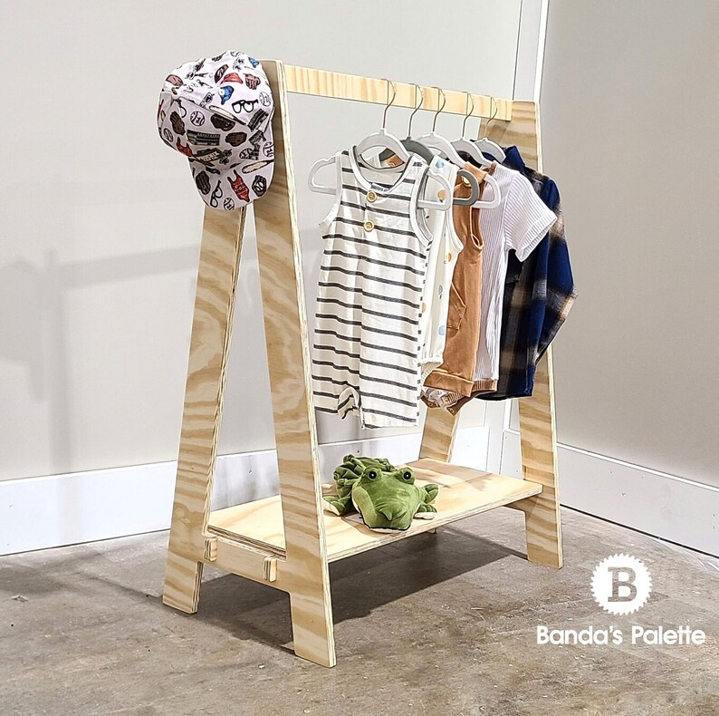 May include: A-frame wooden clothing rack with a shelf, holding several baby outfits on hangers. A patterned baseball cap hangs on the side. A green stuffed frog sits on the shelf. The rack is made of light-colored wood.