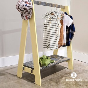 May include: A yellow and gray wooden clothing rack with a shelf, holding children's clothes and a baseball cap. The rack has an A-frame design and a green stuffed frog on the shelf. The clothes include a striped romper and a plaid shirt.