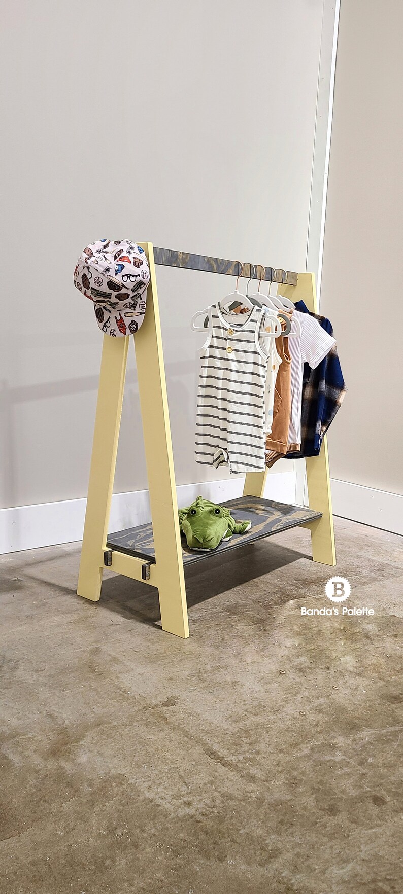 May include: A yellow and gray wooden clothing rack with a shelf, holding children's clothes and a baseball cap. The rack has an A-frame design and a green stuffed frog on the shelf. The clothes include a striped romper and a plaid shirt.