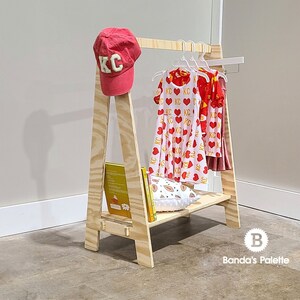 May include: A wooden children's clothing rack with a red KC baseball cap, dresses, and books. The dresses have a heart and KC pattern. The rack has a shelf with a pillow and books. The rack is made of light-colored wood.