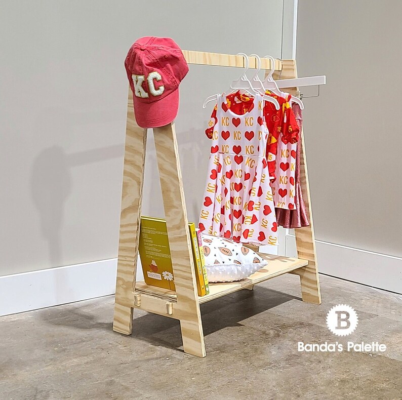 May include: A wooden children's clothing rack with a red KC baseball cap, dresses, and books. The dresses have a heart and KC pattern. The rack has a shelf with a pillow and books. The rack is made of light-colored wood.