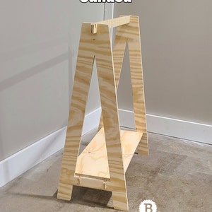 May include: A wooden sawhorse with a natural finish. The sawhorse has an A-frame design with a horizontal shelf. The word "Sanded" is written in white text at the top. The Banda's Palette logo is at the bottom.