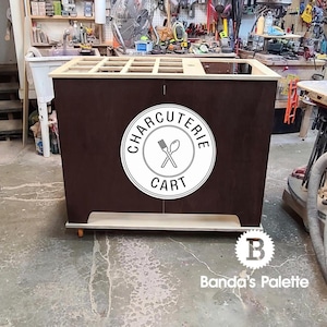 May include: A wooden charcuterie cart with a white circle logo that says "Charcuterie Cart" and a graphic of crossed utensils. The cart has a brown wood finish and a light wood top.