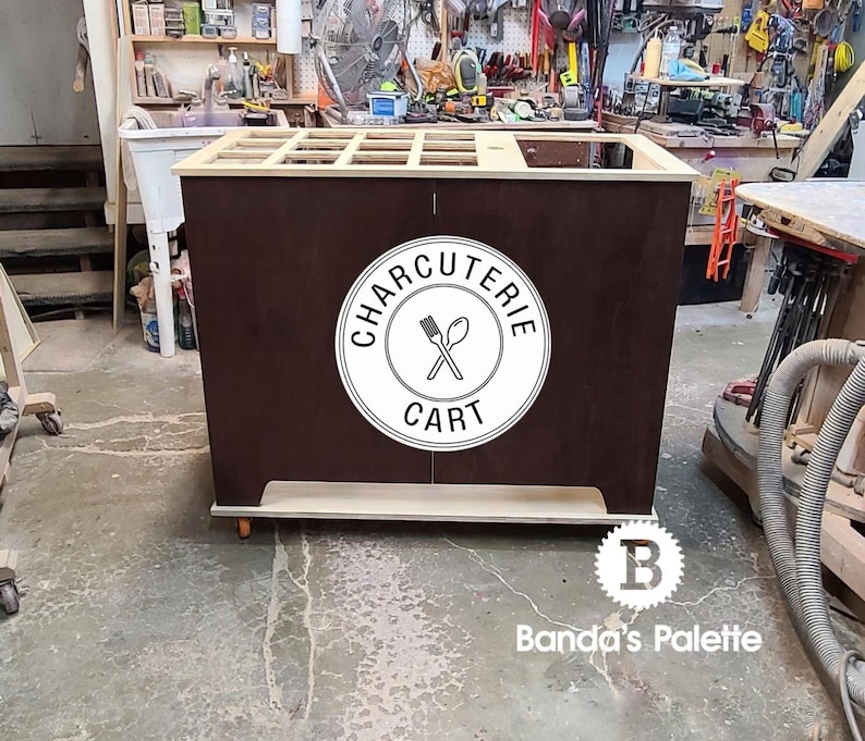 May include: A wooden charcuterie cart with a white circle logo that says "Charcuterie Cart" and a graphic of crossed utensils. The cart has a brown wood finish and a light wood top.