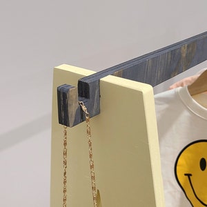May include: A wooden clothing rack with a yellow frame and dark gray accents. A gold chain hangs from the top. A white t-shirt with a yellow smiley face graphic is visible on the rack.