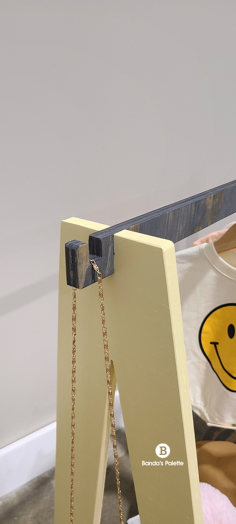 May include: A wooden clothing rack with a yellow frame and dark gray accents. A gold chain hangs from the top. A white t-shirt with a yellow smiley face graphic is visible on the rack.