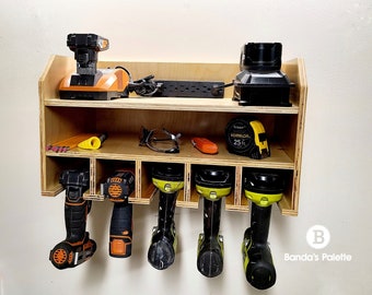 May include: A wooden wall-mounted organizer with six slots for storing power tools. The organizer has a top shelf for storing other items, such as a power cord, a tape measure, and safety glasses. The organizer is made of light brown wood and has a black and orange power tool charger on the top shelf.