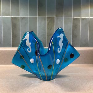 May include: A vibrant, blue glass bowl with a unique, folded design. The bowl features white seahorse designs, colorful dots, and orange and yellow stripes. This decorative piece is a striking addition to any home decor.