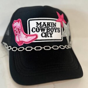 May include: Black trucker hat with a mesh back and a curved brim. The front features a white rectangle with the text "MAKIN COWBOYS CRY". A pink cowboy boot and star are embroidered on the hat, along with a silver chain.