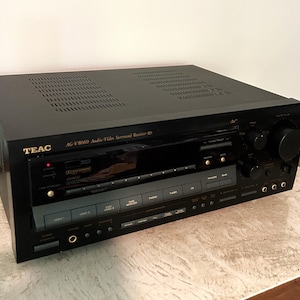May include: Black TEAC AG-V8060 audio/video surround receiver. The front panel has various buttons, dials, and a display screen. The top of the receiver has ventilation holes. The receiver is on a light-colored surface.