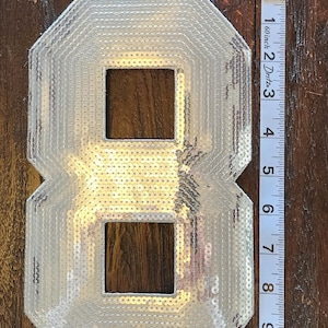 May include: Silver sequined number eight patch, approximately 8 inches tall, with a geometric design. The patch is shown on a dark wood surface next to a measuring tape.