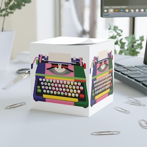 Può includere: Un blocco di foglietti adesivi cubico bianco con una colorata illustrazione grafica di una macchina da scrivere vintage su due lati. Il design della macchina include pannelli viola, verdi, rosa e gialli con una tastiera QWERTY.
