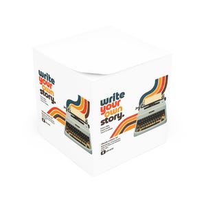 Può includere: Un blocco note cubico bianco con il testo "write your own story." e un'illustrazione grafica di una macchina da scrivere vintage con strisce retrò colorate. Il design include il testo "Olivetti Lettera 82" e il logo Olivetti.