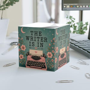 Può includere: Un cubo di foglietti adesivi quadrato con sfondo verde acqua, un'illustrazione di una macchina da scrivere rosa vintage, fiori rosa, una luna crescente e il testo "THE WRITER IS IN" con un piccolo cuore, stampato sui lati del blocco di carta.