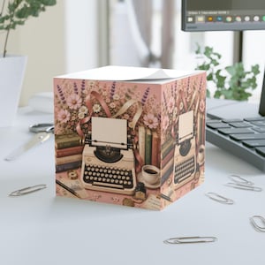 Può includere: Un cubo per appunti quadrato con un'illustrazione stampata di una macchina da scrivere vintage color crema circondata da un bouquet di fiori rosa e bianchi, libri impilati, una tazza di caffè, un calamaio e una penna stilografica su sfondo rosa.