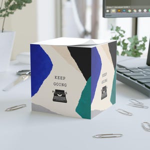 Può includere: Un cubo di foglietti adesivi quadrato con un design a collage di carta strappata in blu, nero, verde acqua e beige. I pannelli frontale e laterale mostrano il testo KEEP GOING sopra un'illustrazione nera di una macchina da scrivere.