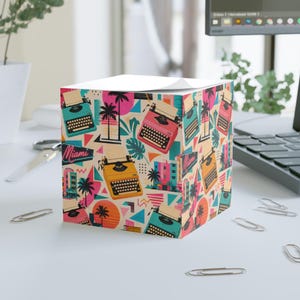 Può includere: Un cubo per appunti quadrato con un motivo ripetuto di macchine da scrivere vintage in verde acqua, rosa e giallo, insieme a palme, forme geometriche e la parola Miami in un carattere in stile neon su uno sfondo color crema.