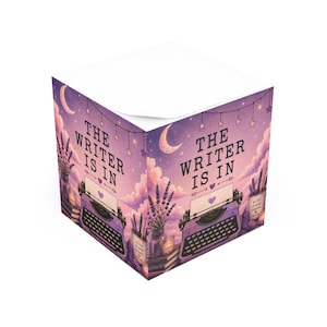 Può includere: Un cubo per appunti quadrato con un'illustrazione viola di una macchina da scrivere vintage, rametti di lavanda e una luna crescente. Il testo THE WRITER IS IN è stampato in nero. Un piccolo contenitore con l'etichetta write dream repeat è accanto.
