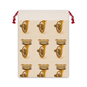 May include: A beige drawstring bag featuring a repeating pattern of vintage brass French horns and typewriters. The instruments have a gold finish and are adorned with small red floral accents. The bag has red drawstrings.