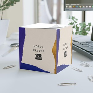 Può includere: Un cubo di foglietti adesivi quadrato con un pannello centrale color crema che riporta il testo nero "WORDS MATTER" e una piccola icona di macchina da scrivere. I lati sono incorniciati da bordi di carta strappata blu e color blu e oro.