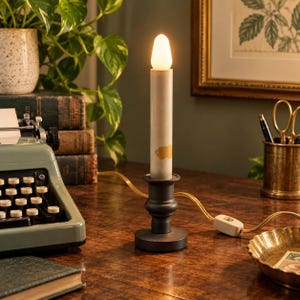 May include: A battery-operated electric candle lamp with a matte gray cylindrical body featuring a gold-colored accent. The lamp sits on a dark, rounded pedestal base and includes a warm-toned, flame-shaped bulb at the top with a power cord.