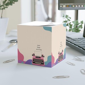 Può includere: Un cubo di foglietti adesivi quadrato con sfondo color crema e forme astratte rosa, blu e viola. Il cubo presenta la grafica di una macchina da scrivere rosa con il testo "just keep writing" stampato sopra su due lati visibili.