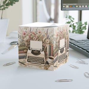 Può includere: Un cubo di foglietti adesivi quadrato con un'illustrazione stampata di una macchina da scrivere vintage color crema circondata da fiori di campo colorati, libri impilati, una penna stilografica, un calamaio e una tazza di caffè.