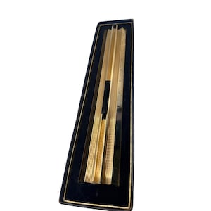 Puede incluir: Una regla de cálculo de metal dorado con una serie de ranuras verticales paralelas y un cursor deslizante negro central, alojada dentro de un estuche rectangular negro con borde dorado a lo largo de los bordes internos del marco.
