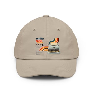 May include: A beige baseball cap featuring a graphic print of a light blue typewriter with colourful retro stripes. The text reads write your own story along with smaller text and a social media icon. The cap has a curved brim and eyelets.