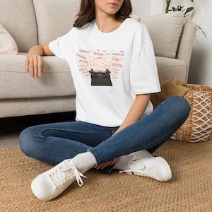 May include: A white short-sleeve t-shirt featuring a graphic of a black vintage typewriter surrounded by red text including words like CREATE, hope, freedom, dream, imagine, life, and story, paired with blue denim jeans and white sneakers.