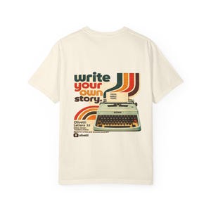 May include: A cream-colored short-sleeve t-shirt featuring a graphic of a light green Olivetti Lettera 32 typewriter. The text reads "write your own story." and "IDEAS. WORDS. YOURS." along with "Olivetti Lettera 32" and "Made for writers and dreamers since 1971."