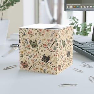Può includere: Un cubo di foglietti adesivi quadrato con sfondo color crema e un motivo ripetuto di macchine da scrivere vintage, penne stilografiche, buste e illustrazioni floreali colorate. Il design include piccoli uccelli ed elementi di testo in corsivo.