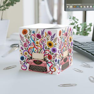 Puede incluir: Un bloque de notas cuadrado de papel con una ilustración de una máquina de escribir rosa en las caras frontal y lateral. El diseño incluye flores coloridas y cintas que se arremolinan saliendo de la máquina sobre un fondo blanco.