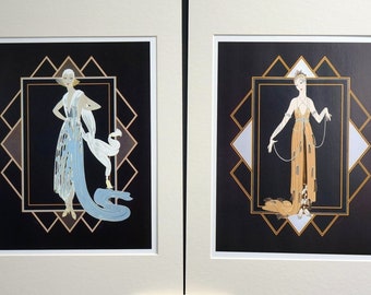 May include: Two framed prints of women in Art Deco style dresses. The woman on the left is wearing a light blue dress with a white feathered shawl and a white bird. The woman on the right is wearing a gold dress with a long beaded necklace. Both prints have a black background with gold and silver geometric designs.