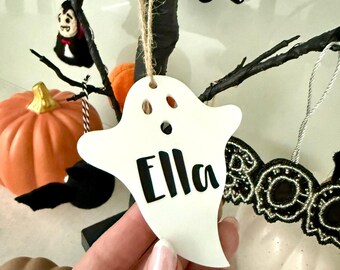 May include: A white ghost-shaped ornament made of a smooth material, featuring the name Ella printed in black lettering. The ornament includes a hole at the top with a brown twine string attached for hanging, designed as a decorative piece.